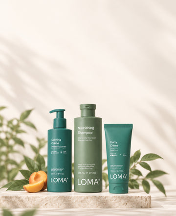 LOMA Calming Crème, Nourishing Shampoo, and Curvy Crème on a stone platform with fruit and green leaves