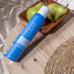 Loma Dry Shampoo 4.4 oz blue aerosol can displayed diagonally on a wooden surface beside pears