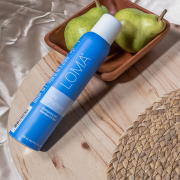Loma Dry Shampoo 4.4 oz blue aerosol can displayed diagonally on a wooden surface beside pears