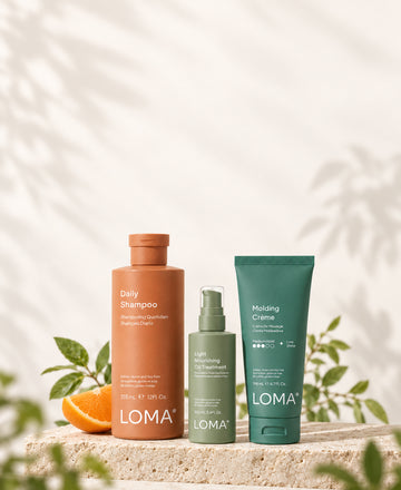 LOMA Daily Shampoo, Light Nourishing Oil Treatment, and Molding Crème on a stone platform with orange and green leaves