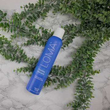 Loma Finishing Hairspray blue aerosol can arranged diagonally on a marble surface with eucalyptus foliage