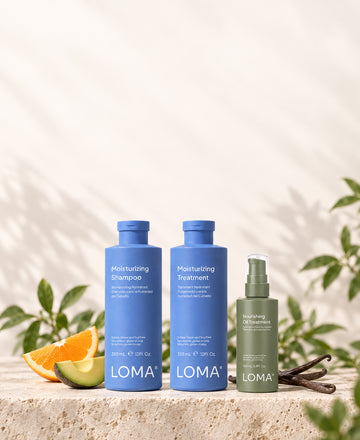 LOMA Moisturizing Shampoo, Moisturizing Treatment, and Nourishing Oil Treatment on a stone platform with orange, avocado, and vanilla pods