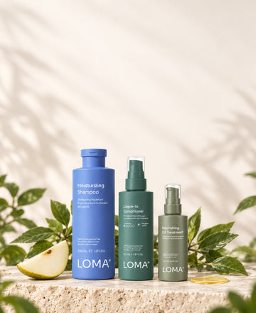 LOMA Moisturizing Shampoo, Leave-In Conditioner, and Nourishing Oil Treatment on a stone platform with pear and green leaves