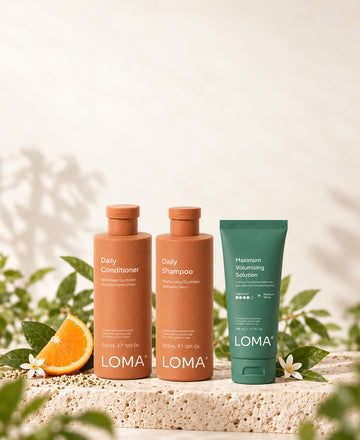LOMA Daily Shampoo, Daily Conditioner, and Maximum Volumizing Solution on a stone platform with citrus slices and green leaves